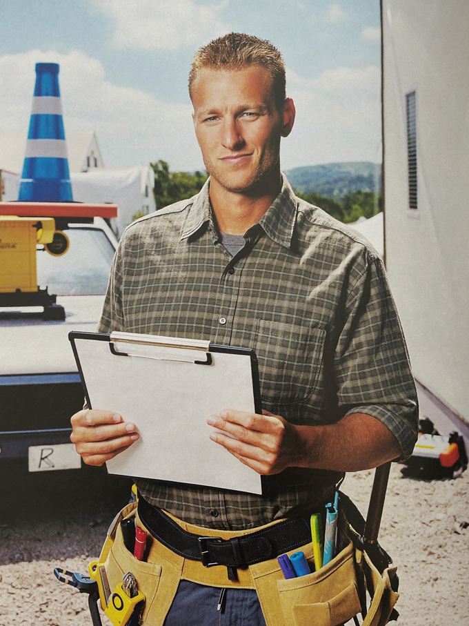 A contractor in a plaid shirt and tool belt holding a clipboard at a work site.