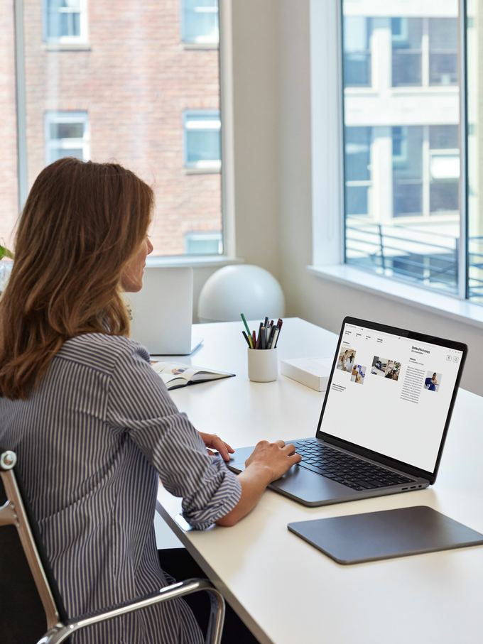 A freelancer working on a laptop at a desk beside tall city windows.